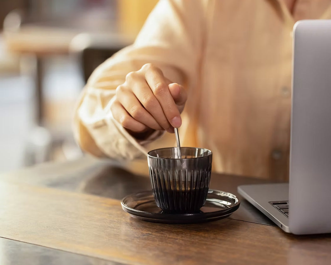 Person stirring espresso in a smoke HuskeeRenew espresso cup and saucer whilst working on laptop in cafe