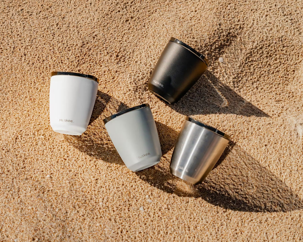 Four HuskeeSteel coffee cups in the sand in black, slate, white, and brushed steel