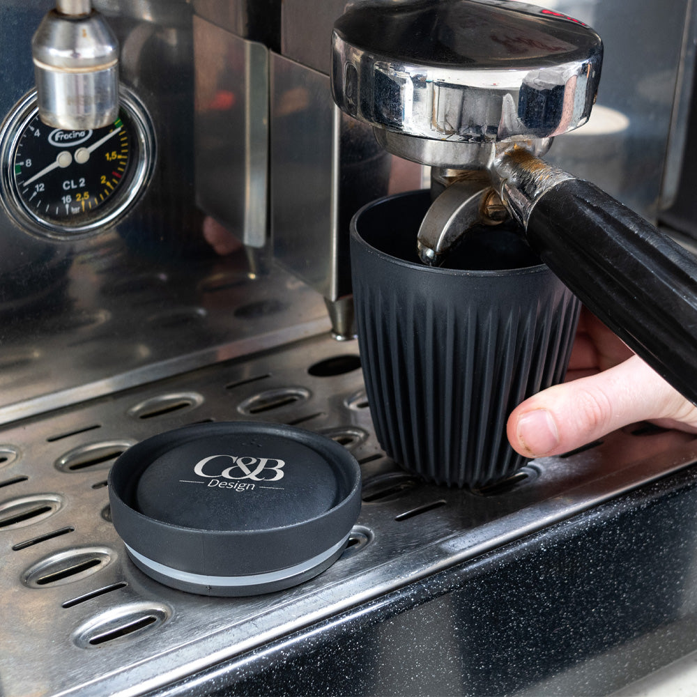Charcoal original coffee cup under the grouphead in an espresso machine with lid next to it with logo on it.